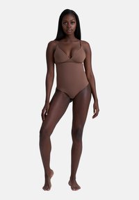 Una donna di pelle scura in piedi a piedi nudi, che indossa un costume da bagno intero marrone con spalline sottili, su uno sfondo chiaro e semplice.