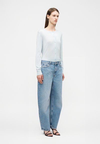 Kvinne iført lyseblå knappet cardigan, høytlivs løse blå jeans og svarte sandaler med hæl og stropper, stående foran en ensfarget hvit bakgrunn.