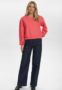 Korallszínű pulóver kerek nyakkal és lejjebb húzott vállakkal, amelyet sötét farmer széles szárú nadrággal és fekete, studdal díszített cipővel párosítottak.