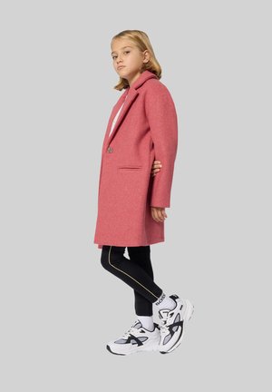 Een jong meisje met blond haar in een roze jas, zwarte leggings met een gele streep, witte "Boss" sokken en zwart-witte sneakers die zijwaarts staat.