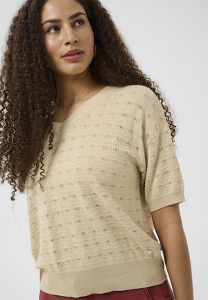 Femme aux longs cheveux bouclés portant un pull court en maille beige et un pantalon bordeaux, regardant légèrement vers le haut à gauche.