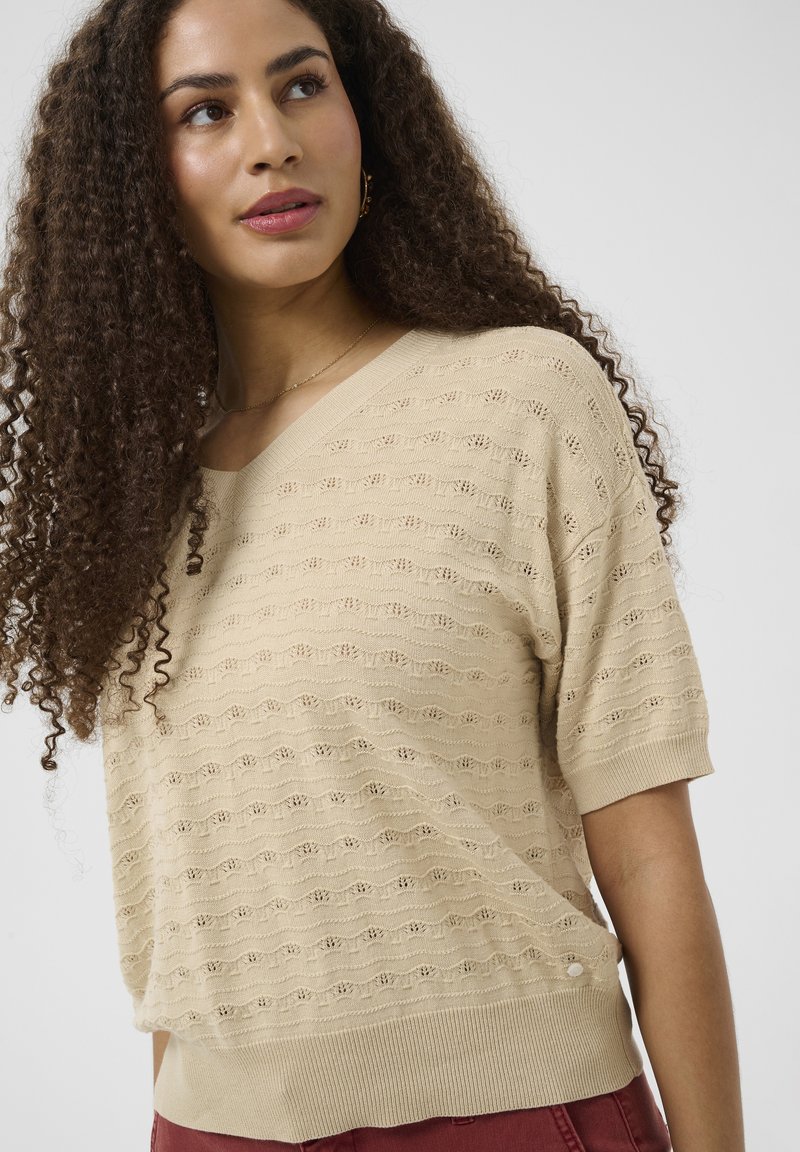 Femme aux longs cheveux bouclés portant un pull court en maille beige et un pantalon bordeaux, regardant légèrement vers le haut à gauche.