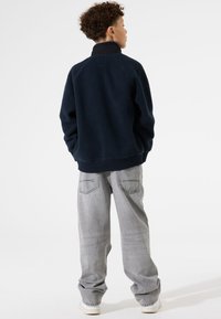 Chaqueta de forro polar azul marino con un alto cuello, que presenta una textura suave y puños acanalados, combinada con unos jeans sueltos de color gris claro.