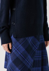 Maglione in maglia blu navy con polsini e orlo a coste, caratterizzato da due bottoni sui lati. Abbinato a una gonna di lana a quadri blu.