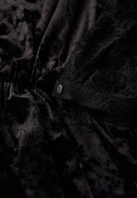 Tela de terciopelo negro con una textura suave que presenta un acento de encaje y un detalle de botón a presión metálico. El diseño general es elegante y ajustado.