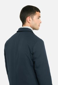 Giacca navy dal design sartoriale, con colletto a labbro, texture liscia e silhouette snella. La vista posteriore mostra cuciture sottili.