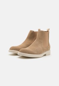 Par de botas Chelsea de ante marrón claro con paneles laterales elásticos y suelas de goma blanca sobre un fondo blanco.