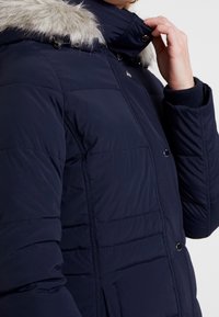 Giacca trapuntata blu navy con cappuccio in pelliccia sintetica removibile, dotata di bottoni a pressione e texture a quadri. Tasche laterali visibili.