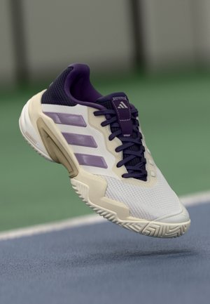 Chaussure de sport Adidas blanche et violette flottant au-dessus d'une surface de terrain de tennis bleue et verte.