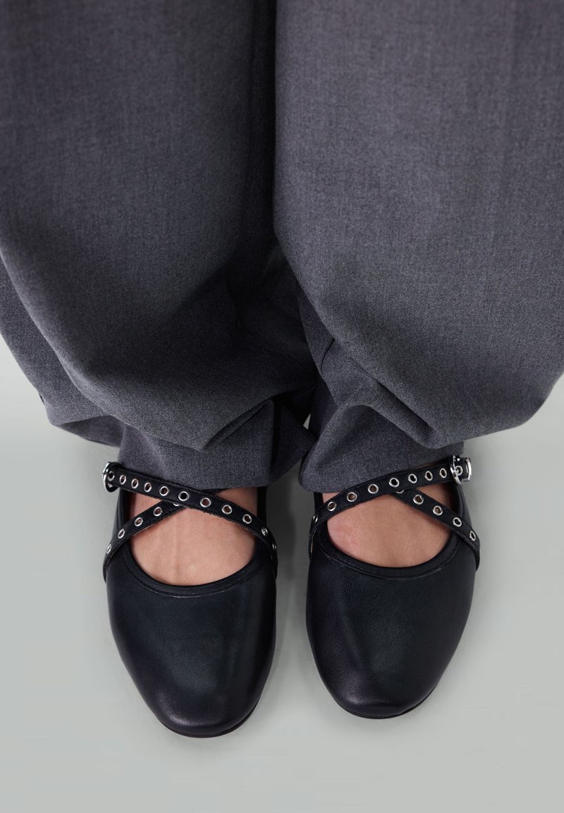 Schwarze Leder-Clogs mit runder Zehenpartie und seitlichen Ausschnitten, versehen mit einem Kreuzriemen, der mit silbernen Ösen verziert ist, getragen mit grauen Hosen.