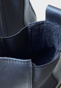 Schwarze Lederstiefelette mit glatter Oberfläche, die ein geflochtenes Stofffutter aufweist. Das Innere zeigt eine robuste Struktur mit verstärkten Kanten.