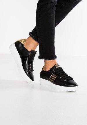 Osoba nosząca czarne trampki ze złotymi akcentami i białymi grubymi podeszwami, w połączeniu z czarnymi spodniami z postrzępionymi brzegami.