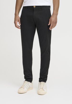 Black, slim-fit pants made of cotton with a button closure and front pockets; paired with off-white sneakers featuring three stripes.