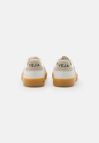 Hvite lær sneakers med beige semsket hællapp, gummisåle og "VEJA" trykt på hælen. Teksturerte overflater med glatte accenter.