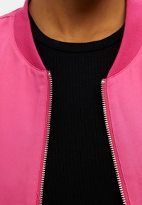 Giacca bomber fucsia con collo a costine e dettagli con cerniera argentata, indossata sopra una maglia nera a costine. Il tessuto sembra liscio e leggero.