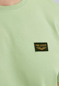 Camiseta de algodón verde claro con cuello redondo. Presenta un parche negro rectangular con texto amarillo y un gráfico de alas en el pecho.