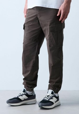 Mann trägt dunkelbraune Cargohosen und schwarz-weiße Sneakers und steht auf einem hellen Boden mit einfachem Hintergrund.