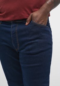 Jeans din denim de culoare albastră închisă, cu cusături portocalii. Prezintă o buzunar frontal și un design ajustat. Materialul pare neted cu o ușoară textură.