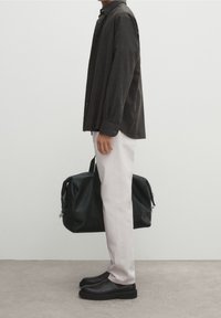 Czarna koszula z długim rękawem, jasne spodnie, czarne buty i czarna torba sportowa; stojąca na teksturowanej betonowej nawierzchni na tle białym.