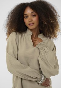 Világos bézs, könnyű blúz magas nyakkal és puffos ujjakkal. Ráncolt mandzsettával és laza fazonnal rendelkezik, texturált anyagból készült.