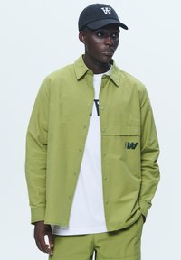 Green button-up shirt with a chest pocket and logo detail. Paired with a white t-shirt and matching green pants, topped with a black cap.