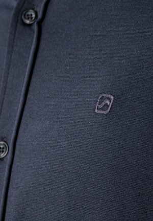 Tumesinine nööpidega särk, mille rinnal on väike tikitud ruudukujuline logo, millel on kõverjoon.