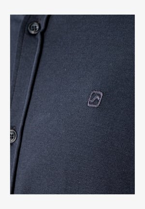 Tumesinine nööpidega särk, mille rinnal on väike tikitud ruudukujuline logo, millel on kõverjoon.