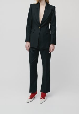 Femme portant un tailleur noir ajusté avec des boutons dorés, des chaussettes rouges et des escarpins blancs à bout pointu devant un fond clair.