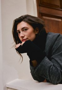 Model nosi tamnosivi teksturirani sako s crnim rolkom i rukavicama bez prstiju, naslonjen na svijetli zid, s drvenim vratima u pozadini.