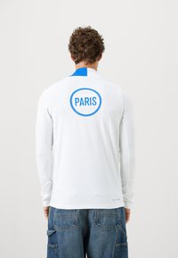 Langærmet hvid skjorte med blå detaljer, der har et cirkulært "PARIS" grafik på ryggen. Glat tekstur med subtile riller.