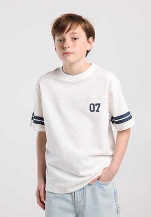 Jeune personne aux cheveux bruns courts, portant un t-shirt blanc avec des rayures bleu marine sur les manches et le numéro 07, jean bleu clair, debout avec une main dans la poche.