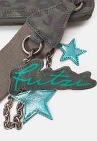 Zielony materiał w cętki przypominające lamparcie plamy z szarym paskiem, z metaliczną, turkusową gwiazdką i czarne etykietą z napisem "fritzi" w turkusowym kursywie. Zawiera szczegóły łańcuchowe.