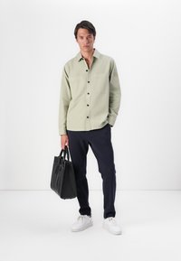 Lichtgroen overhemd met knopen en twee borstzakken, gecombineerd met marineblauwe broek en witte sneakers, terwijl een zwarte tote bag wordt vastgehouden. Minimalistisch ontwerp.