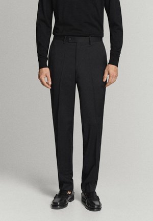 Hombre vistiendo pantalones de vestir negros plisados, camiseta negra de manga larga y mocasines de cuero negro, de pie sobre un fondo gris claro.