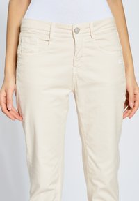 Pantalones beiges de algodón con un diseño ajustado, bolsillos frontales y un pequeño parche con el logo. Presentan un cierre de cremallera y botón.