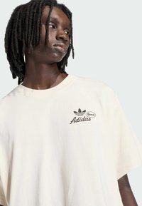 Oversized tričko v světle krémové barvě z bavlny s tmavě hnědým logem Adidas a listovým emblémem na levé straně hrudi. Krátké rukávy.