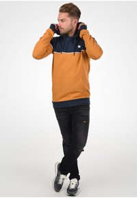 Orangenes und marineblaues Kapuzenpullover mit einer Kängurutasche, ausgestattet mit einem Farbblockdesign und weißen Akzenten. Getragen mit verwaschenen schwarzen Jeans und Sneakers.