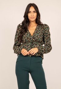 Blouse florale avec un fond sombre, manches bouffantes et taille froncée, associée à un pantalon vert taille haute.