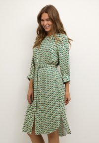 Rochie verde cu un model triunghiular, dintr-un material ușor, având mâneci lungi, brâu la talie și o fanta frontală pentru un plus de libertate de mișcare.