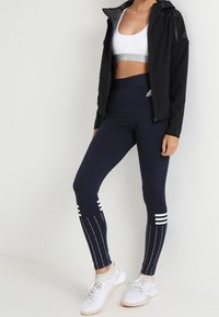Kobieta ubrana w czarną kurtkę zapinaną na zamek, białą biustonosz sportowy, granatowe legginsy Adidas z białymi paskami i białe buty sportowe, stojąca na tle jednolitym.