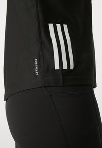T-shirt de sport noir avec texture en maille, arborant trois rayures blanches horizontales et une étiquette "AEROREADY" sur le côté. Vue rapprochée.