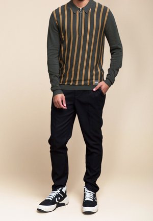 Homme portant un pull à manches longues vert foncé avec fermeture éclair et rayures verticales jaunes, pantalon noir et baskets noir et blanc, debout avec une main dans la poche.