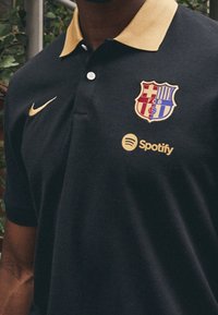 Námorní modré polo tričko s zlatým límcem, logem Nike, znakem FC Barcelona a logem Spotify, s hladkým povrchem a klasickým střihem.