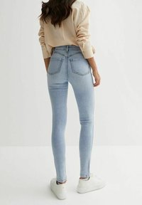 Tight blå denimjeans med hög midja, kontrasterande sömmar och två bakfickor. Matchas med vita sneakers på en slät yta.