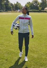 Dres Adidas w jasnoniebieskim kolorze z różowymi akcentami, czarne spodnie i piłka nożna w czarno-białym wzorze. Tło z zielonego boiska.