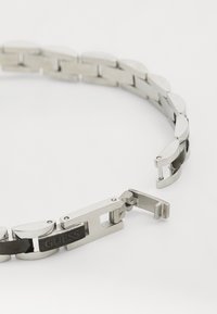 Sidabrinė metalinė apyrankė su ovaliais žiedais, turinti juodą sekciją ir išgraviruotą logotipą. Matoma sagties mechanizmas, rodantis poliruotą paviršių.