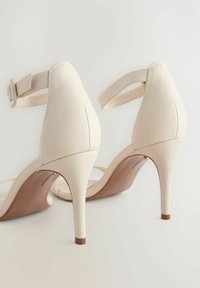 Sandalias de tacón alto de piel blanca con acabado texturizado, correa en el tobillo y tacón de aguja afinado; punta abierta y plantilla beige.