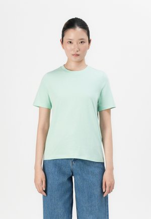 Mujer con el pelo negro atado hacia atrás, vistiendo una camiseta de manga corta de color verde claro y jeans azules, de pie frente a un fondo blanco.