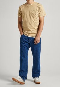 Beige bomullströja med uppvikta ärmar och broderad logotyp, ihop med lösa blå denimjeans och vita sneakers med beige sula.