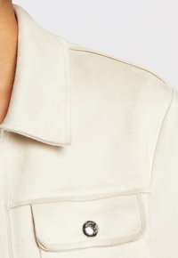 Beige suède shirt met een kraag, een enkele borstzak en zilveren hardwareaccenten. Zachte textuur met een gladde afwerking.
